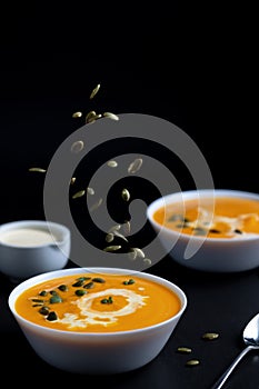 Yellow cream soup in the white dish on the black table and falling seeds