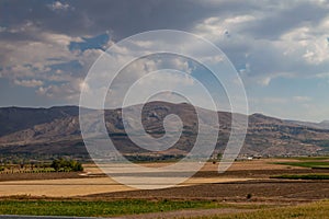 Yellow dry valley with freshly harvested hay bales, other green fields, mountains and blue sky. Dots of dark green tree foliage.