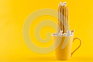 A yellow ear of corn sticking out of a yellow cup, as if growing, popcorn is scattered, on a yellow background, concept