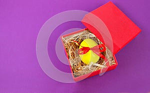 A yellow Easter egg tied with a red ribbon lies in a red gift box on a gray background.