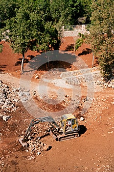 Yellow excavator works on a red quarry against a background of green trees