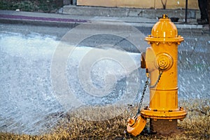 Yellow fire hydrant wide open gushing water onto the street with slightly grainy effect where water is falling back down over the