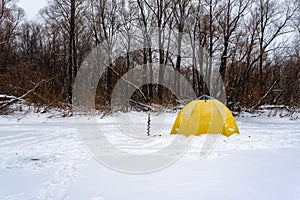 Yellow fishing winter tent stands with an ice-hole in the winter on the ice near the edge of the forest on a snow storm