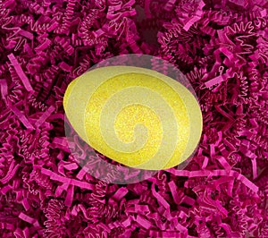 Yellow glitter easter egg atop dark pink shredded crinkle paper overhead view