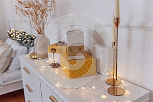 Yellow, gold gift boxes lie on a modern bedside table. The vase contains dried flowers, candlesticks