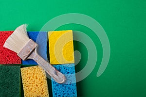 Yellow, green, red, blue sponges with paint brush on green colored paper background, copy space, top view, flat lay