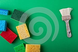 Yellow, green, red, blue sponges with paint brush on green colored paper background, copy space, top view, flat lay