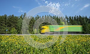Yellow-green truck rushes along the road along the forest, the roadside is covered with yellow dandelions
