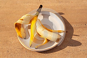 Yellow half peeled banana on the plate on brown background with sharp shadows. Fresh organic fruit on sunny day. Close up. Still