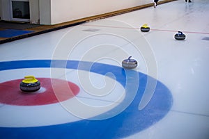 A yellow-handled curling stone sits on the ice in the center of the house, with the other stones out of focus in the back