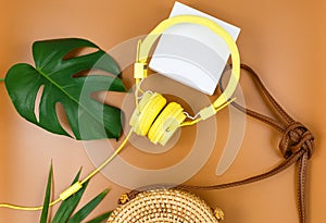 Yellow headphones with tropic leaves, straw bag on brown background. Trendy fashion accessories. Flat lay, close up. Summer,