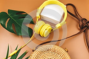 Yellow headphones with tropic leaves, straw bag on brown background. Trendy fashion accessories. Flat lay, close up. Summer,