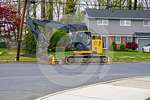 Yellow heavy equipment excavator parked on the side of a residential street in front of a house with orange warning cones