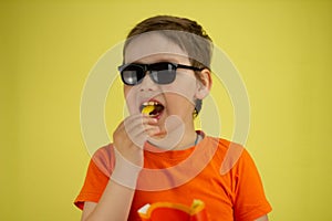 On a yellow isolated background, a child bites off a piece of french fries. A child in sunglasses eats crispy fried