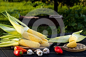 Yellow juicy corn with green leaves lies on a wooden table in the summer garden against the backdrop of a grill