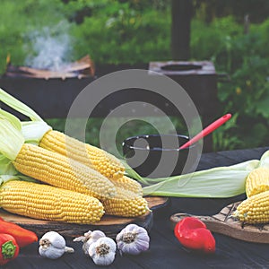 Yellow juicy corn with green leaves lies on a wooden table in the summer garden against the backdrop of a grill