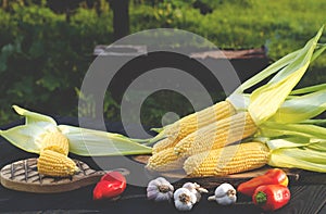 Yellow juicy corn with green leaves lies on a wooden table in the summer garden against the backdrop of a grill