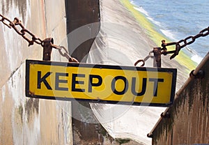 Yellow keep out sign hung on a chain blocking the entrance to concrete sea wall leading to the beach at high tide