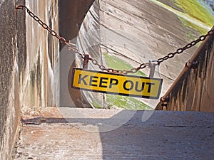 Yellow keep out sign hung on a chain blocking the entrance to concrete sea wall leading to the beach at high tide