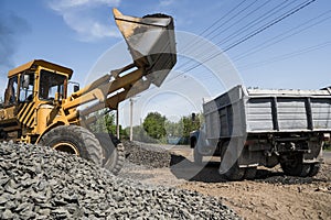Yellow loader delivering stone gravel into truck during road construction works. The stones for the road. Unloading