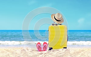 Yellow luggage with hat and flip-flop on sandy beach under blue sky background