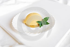 Yellow mousse cake in the form of a lemon lies on a white plate with a spoon on a light background.