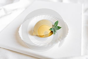 Yellow mousse cake in the form of a lemon lies on a white plate with a spoon on a light background.