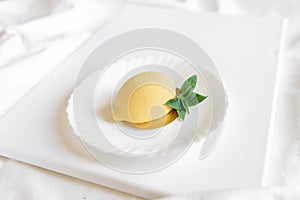 Yellow mousse cake in the form of a lemon lies on a white plate with a spoon on a light background.