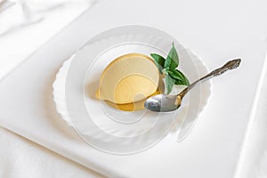 Yellow mousse cake in the form of a lemon lies on a white plate with a spoon on a light background.