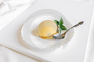 Yellow mousse cake in the form of a lemon lies on a white plate with a spoon on a light background.