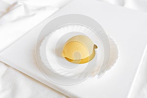 Yellow mousse cake in the form of a lemon lies on a white plate with a spoon on a light background.