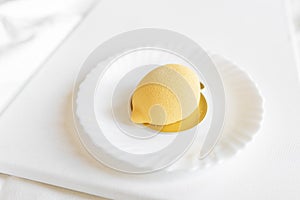 Yellow mousse cake in the form of a lemon lies on a white plate with a spoon on a light background.