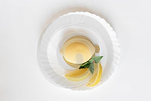 Yellow mousse cake in the form of a lemon lies on a white plate with a spoon on a light background.