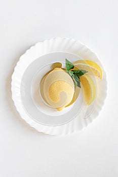 Yellow mousse cake in the form of a lemon lies on a white plate with a spoon on a light background.