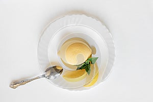 Yellow mousse cake in the form of a lemon lies on a white plate with a spoon on a light background.