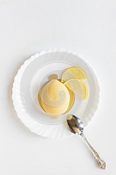 Yellow mousse cake in the form of a lemon lies on a white plate with a spoon on a light background.