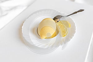 Yellow mousse cake in the form of a lemon lies on a white plate with a spoon on a light background.