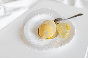 Yellow mousse cake in the form of a lemon lies on a white plate with a spoon on a light background.