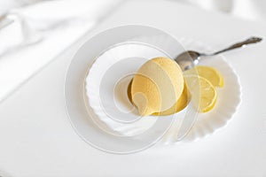 Yellow mousse cake in the form of a lemon lies on a white plate with a spoon on a light background.