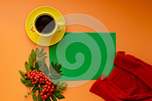 A yellow mug with a saucer, red sweater, and rowan berries on an orange background lay flat. Green sheet of paper copy space