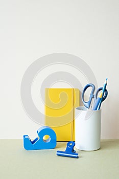 Yellow notebook, blue colored pencil, pen, scissors, clip, tape dispenser on desk. white ivory background. workspace office