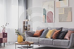 Yellow, orange, black and brown pillows on comfortable grey scandinavian sofa in bright living room interior with abstract