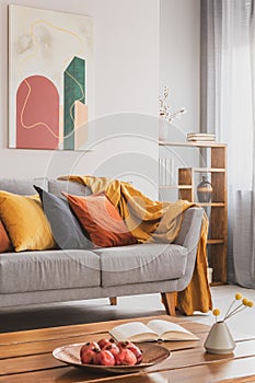 Yellow, orange, black and brown pillows on comfortable grey scandinavian sofa in bright living room interior with abstract