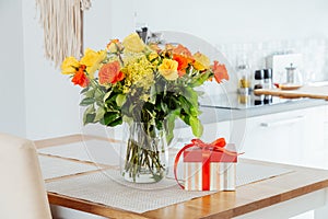 Yellow and orange roses in a vase and gift box with red ribbon on kitchen counter table with modern kitchen