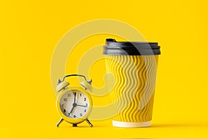 Yellow paper cup of coffee next to yellow alarm clock on a yellow background. Getting up early in the morning concept.