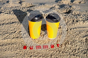 Yellow paper cups with coffee on the sand in the sunset rays of the sun. The inscription SUMMER in red letters