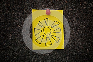 A yellow paper note with a drawing of the sun on it pinned to a cork board