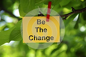 A yellow paper note with the phrase Be The Change on it attached to a tree branch with a clothes pin