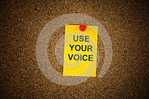 A yellow paper note with the slogan Use Your Voice on it pinned to a cork board