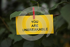 A yellow paper note with the words You Are Unbreakable on it attached to a tree branch with a clothes pin